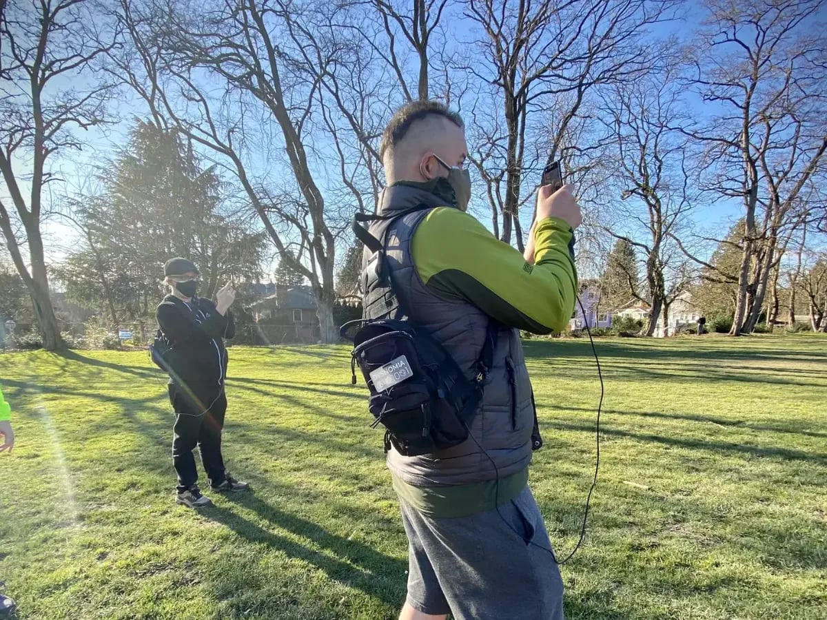 Two researchers standing on a park lawn on a bright winter day, each holding a phone up at eye level, wearing chest rigs with capture gear and cables running to the handsets.