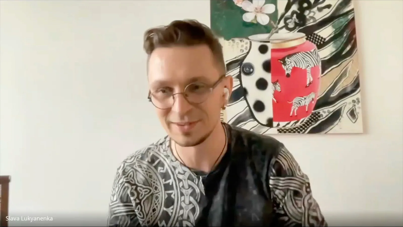 A GSC Game World team member talking to camera on a webcam call in Live Aware, in a room with a patterned wall hanging behind him.